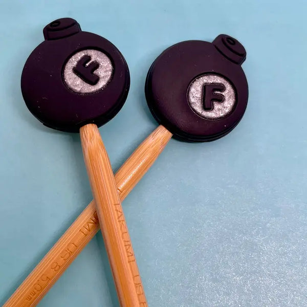 Two black bomb shapes with 'F' on them on the tips of light brown knitting needles on a light blue background