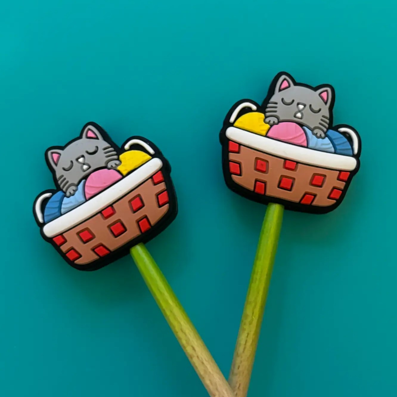 Two silicone needle point protectors in shape of  a brown and red basket with a light grey cat and multicolored yarn peeking out, attached to wooden sticks, against a blue background