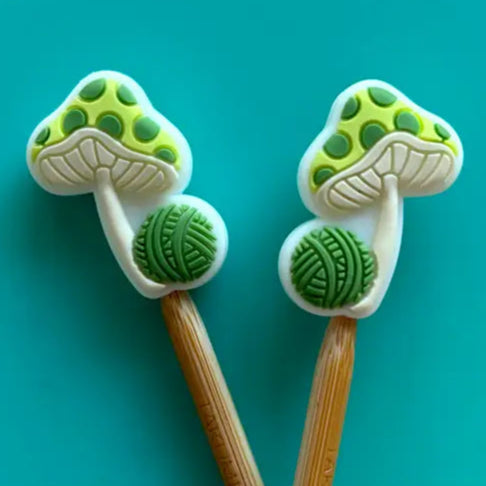 Two wooden knitting needles with green mushroom-shaped ends on a teal background